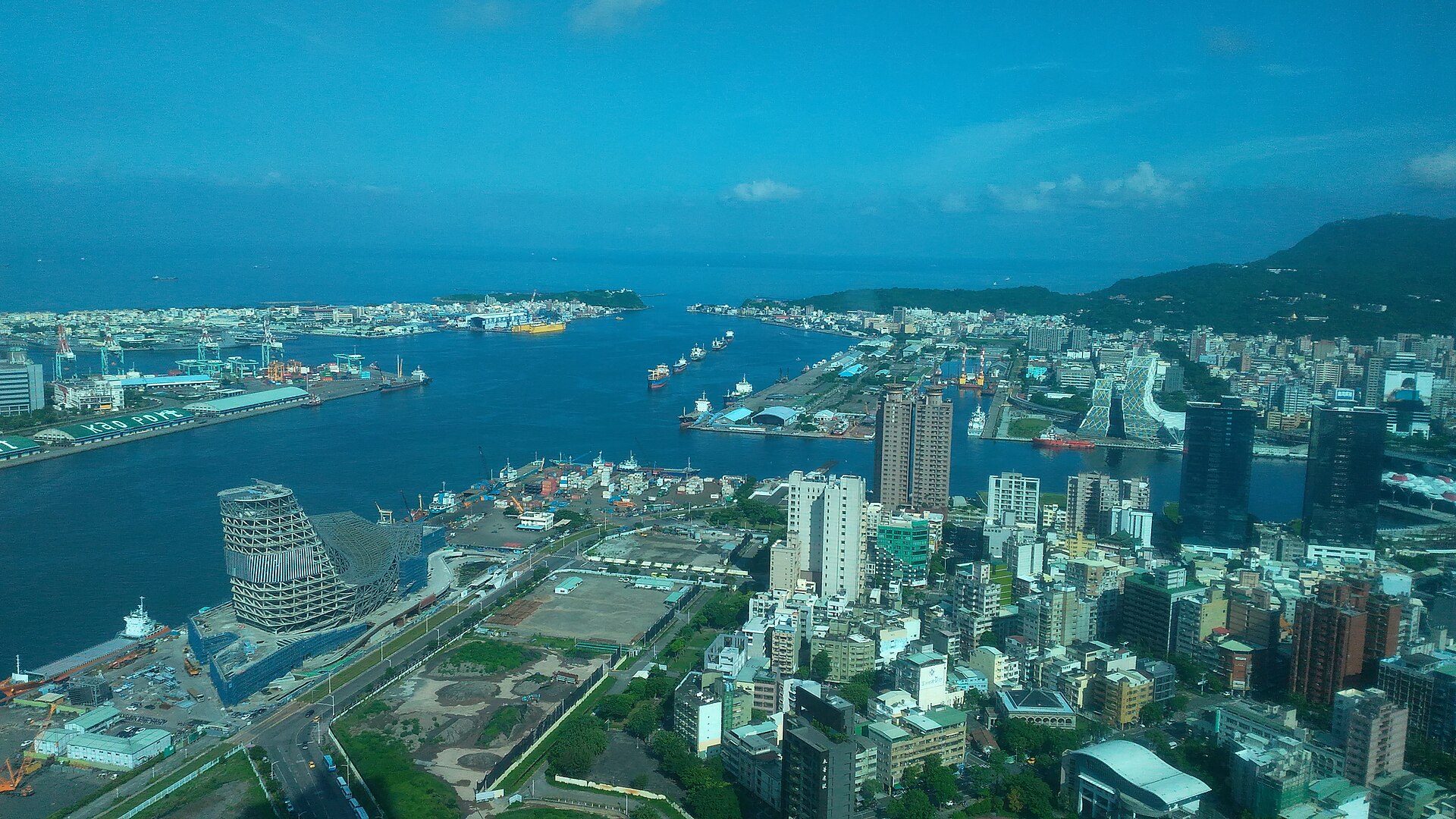 高雄港第一港口空拍,貨櫃碼頭與起重機沿著岸邊排列,貨船停靠在航道兩側,後方是高雄市區與壽山